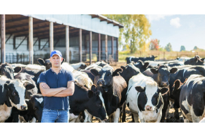A farmer stands outside in front of a herd of black-and-white cows. They are near a steel building on a sunny day.