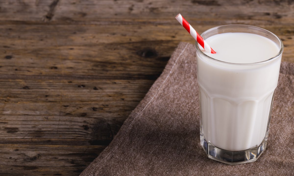 A red and white striped straw is in a full glass of white milk. The glass is on a brown placemat on a wooden table.